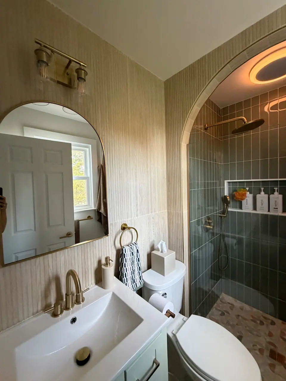 Arched shower entry with sage green vertical tile, brass fixtures and an arched mirror