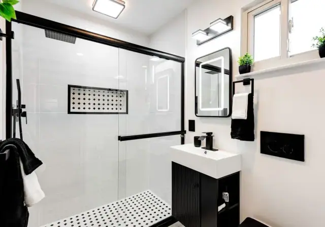 Black and white bathroom with a glass shower, black fixtures and a lit mirror
