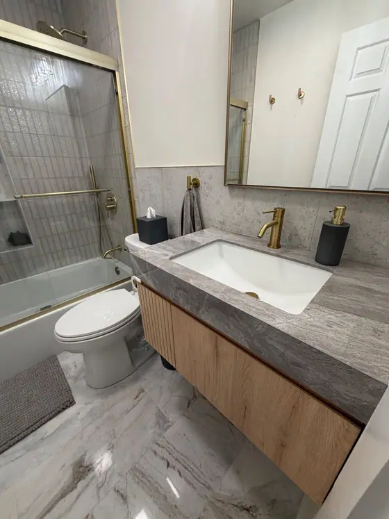 Floating wood vanity with a stone top and brass faucet beside a glass tub enclosure