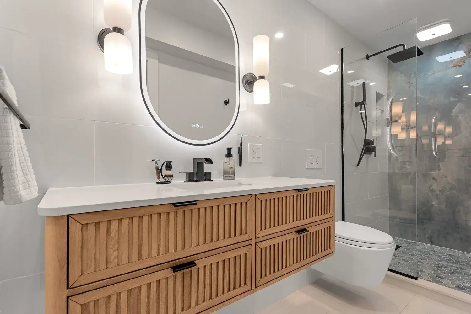 Bathroom with a floating fluted wood vanity, lit oval mirror and a walk in shower with stone look tile