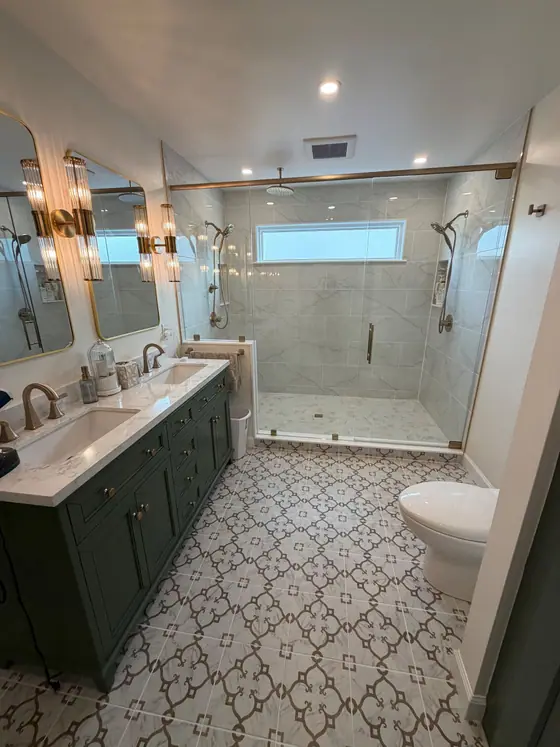Double vanity in dark green with a patterned tile floor leading to a glass shower