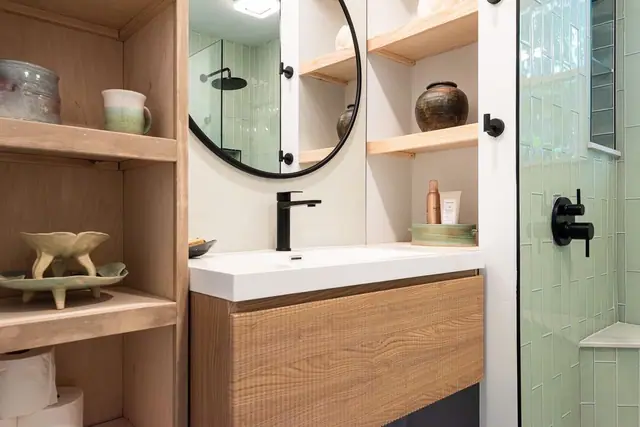 Open wood shelving beside a white vanity, round black mirror and sage green shower tile