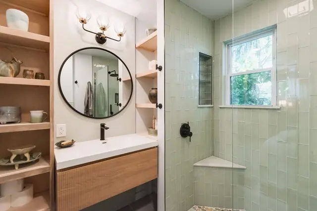 Sage green tile shower with a corner bench next to a wood vanity and open shelves