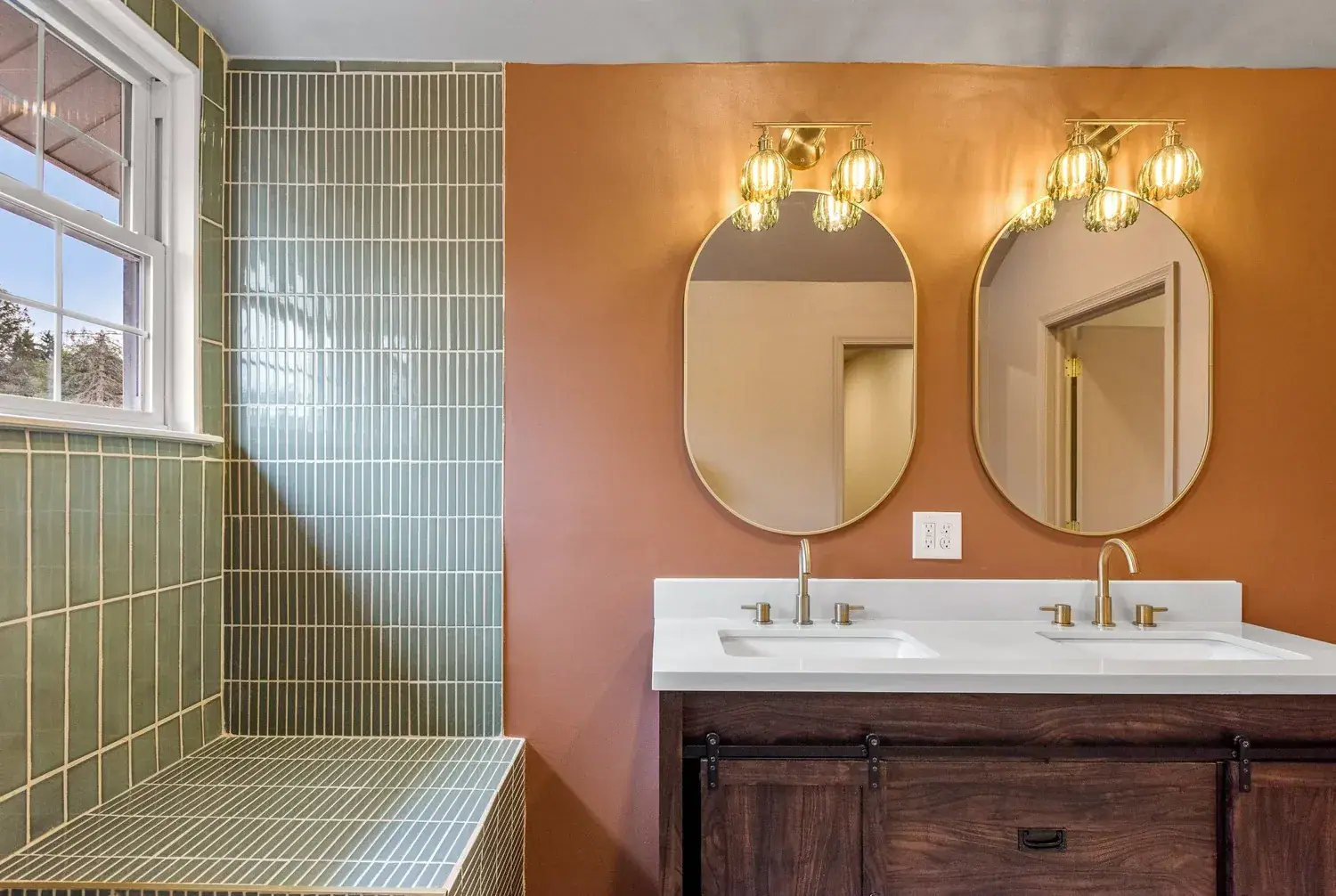 Double vanity bathroom with green stacked tile, terracotta walls and brass pendant sconces