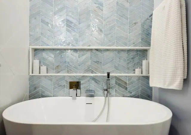 Freestanding soaking tub below a blue chevron tile wall with a long built in niche